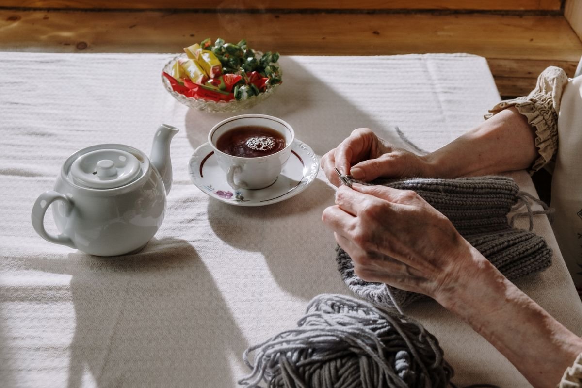 An older woman's hands knitting at her own kitchen table, with tea poured, in afternoon sunlight